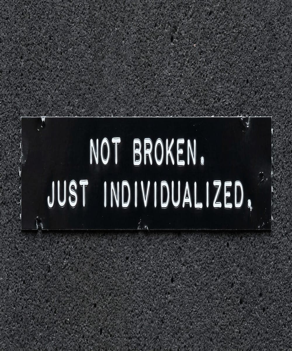 Punched label-tape plaque reading 'Not broken. Just individualized.' screwed to a dark speckled wall.
